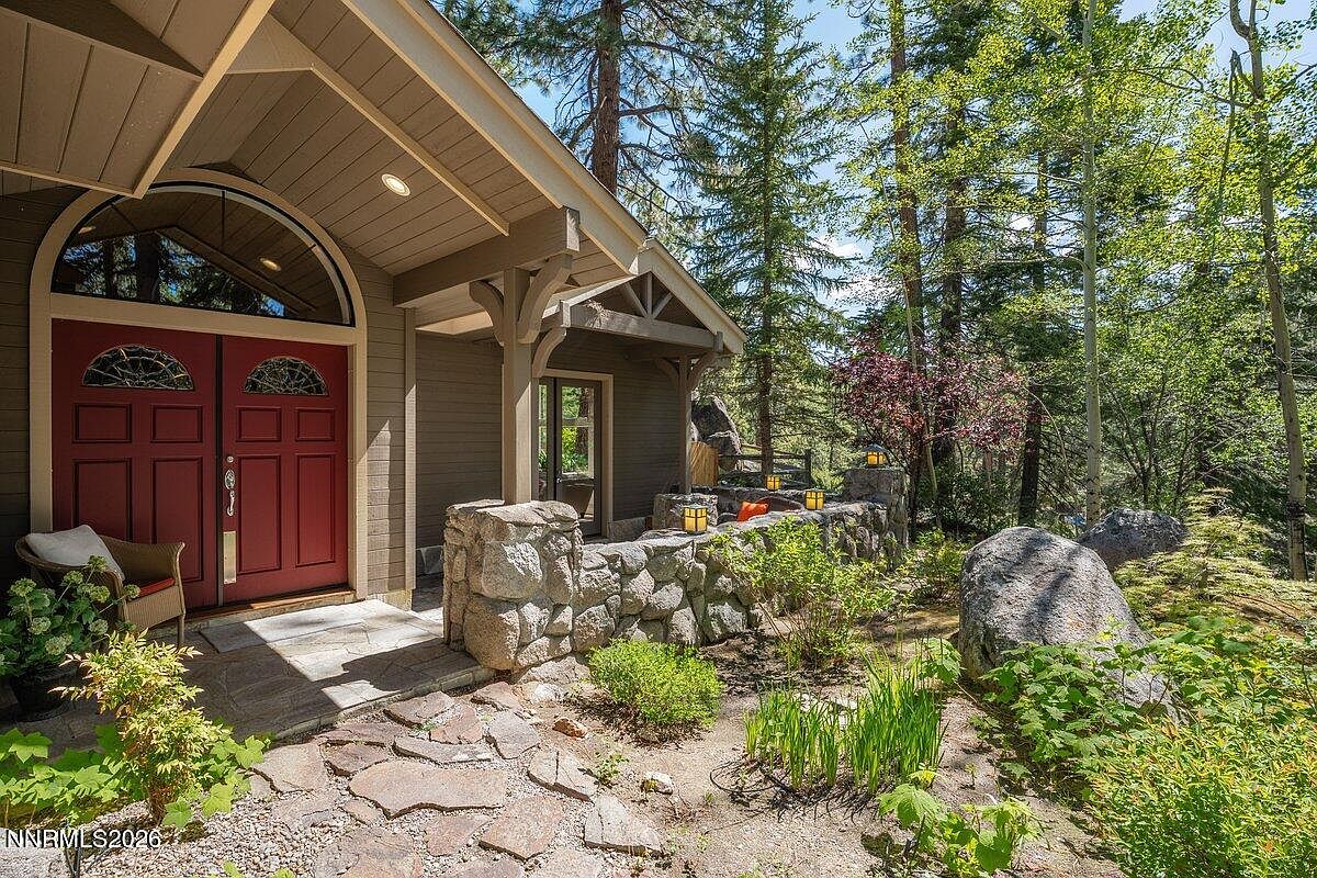 Stone-flanked covered entry to 1 Kelly Cir with red double doors, arched timber porch, and a stone bench set among ferns and towering pines