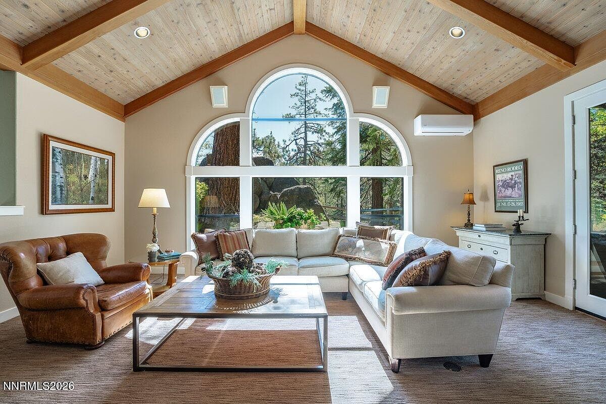 Great room of 1 Kelly Cir with exposed timber beams, vaulted wood ceiling, a large arched window framing granite boulders and pine forest, sectional sofa, leather club chair, and stone fireplace surround
